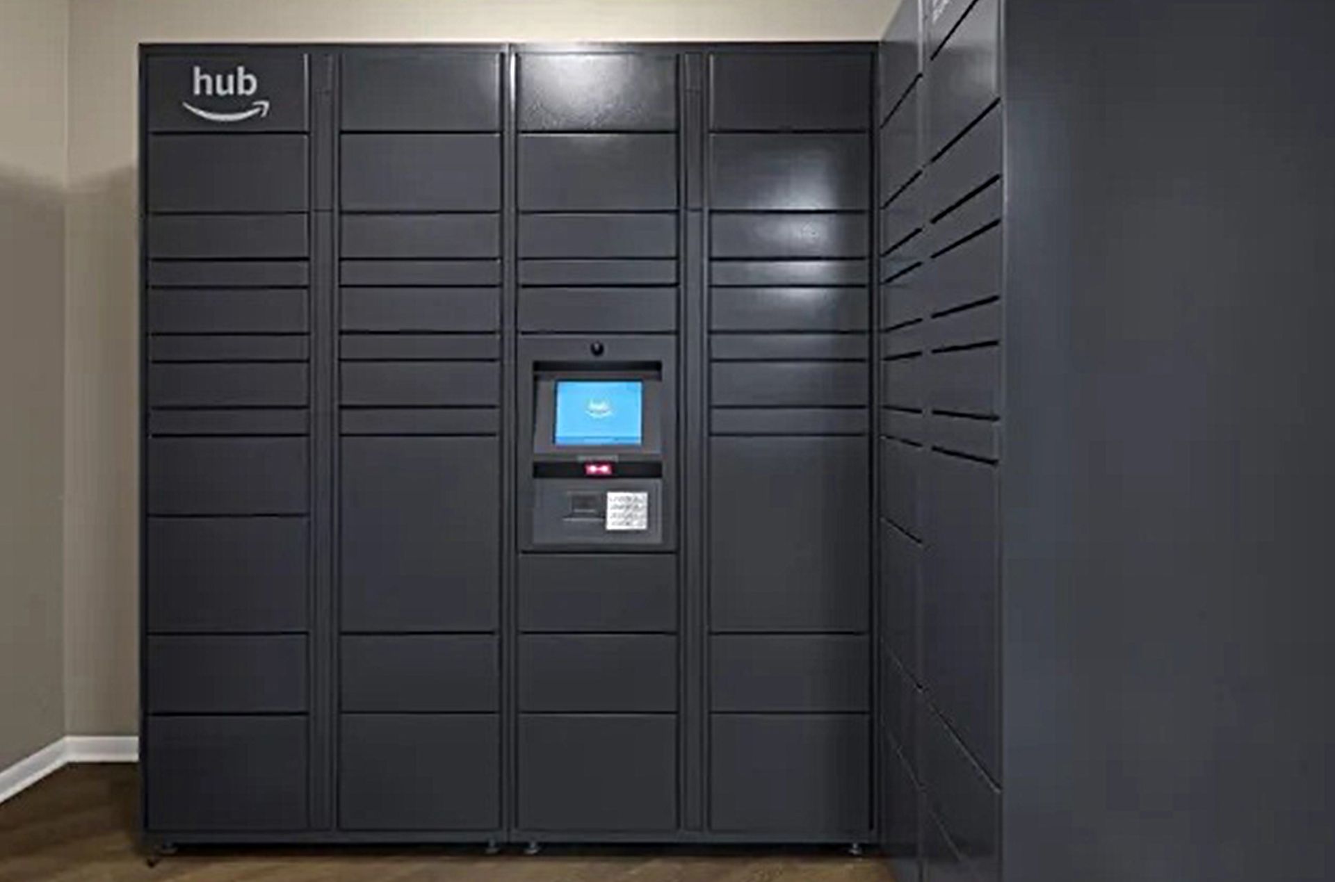 Amazon Hub locker system in a building hallway. Gray metal lockers with a touchscreen.