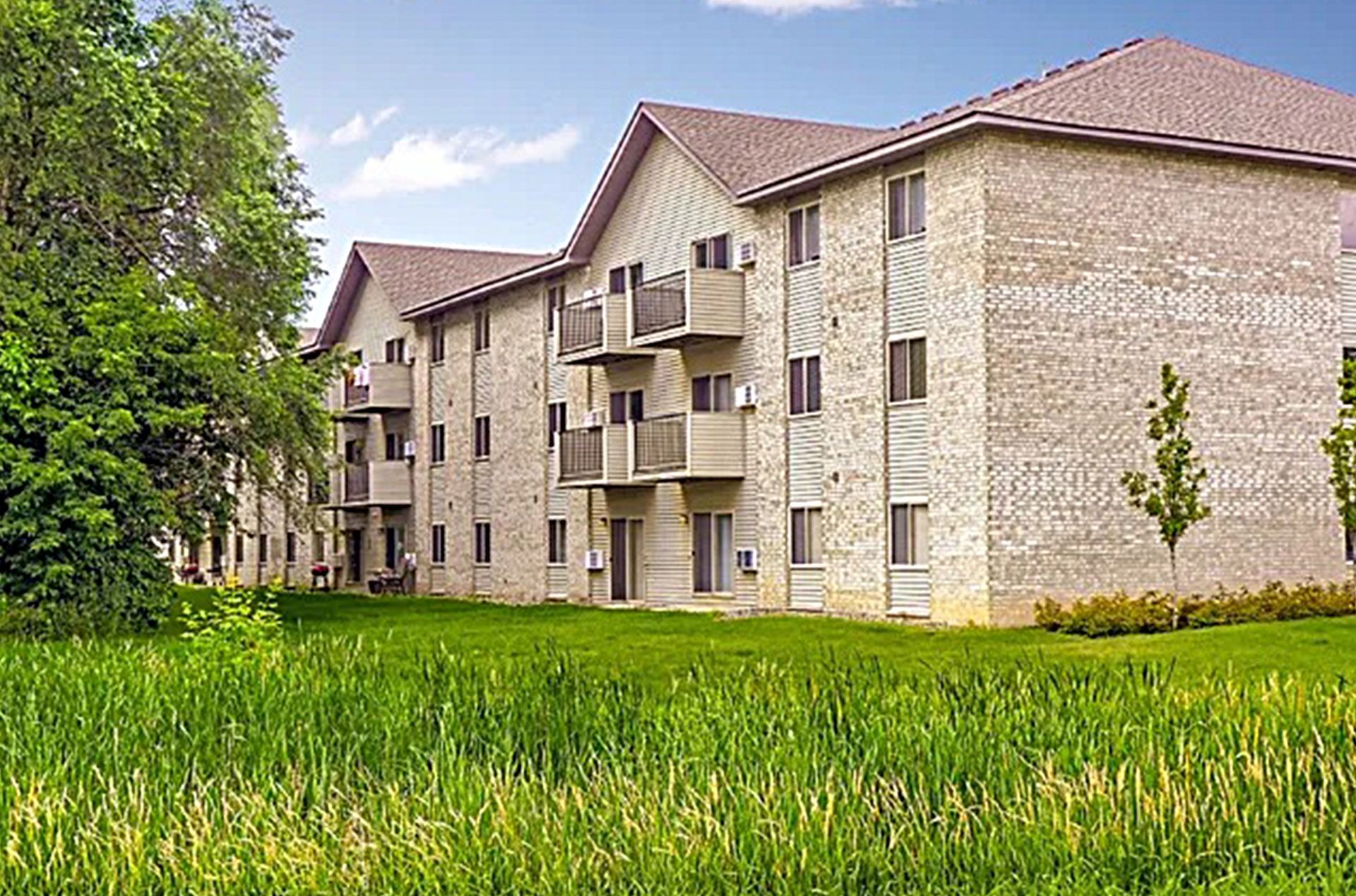 Apartment building with beige brick exterior, brown roof, and balconies, viewed from grassy field on a sunny day.