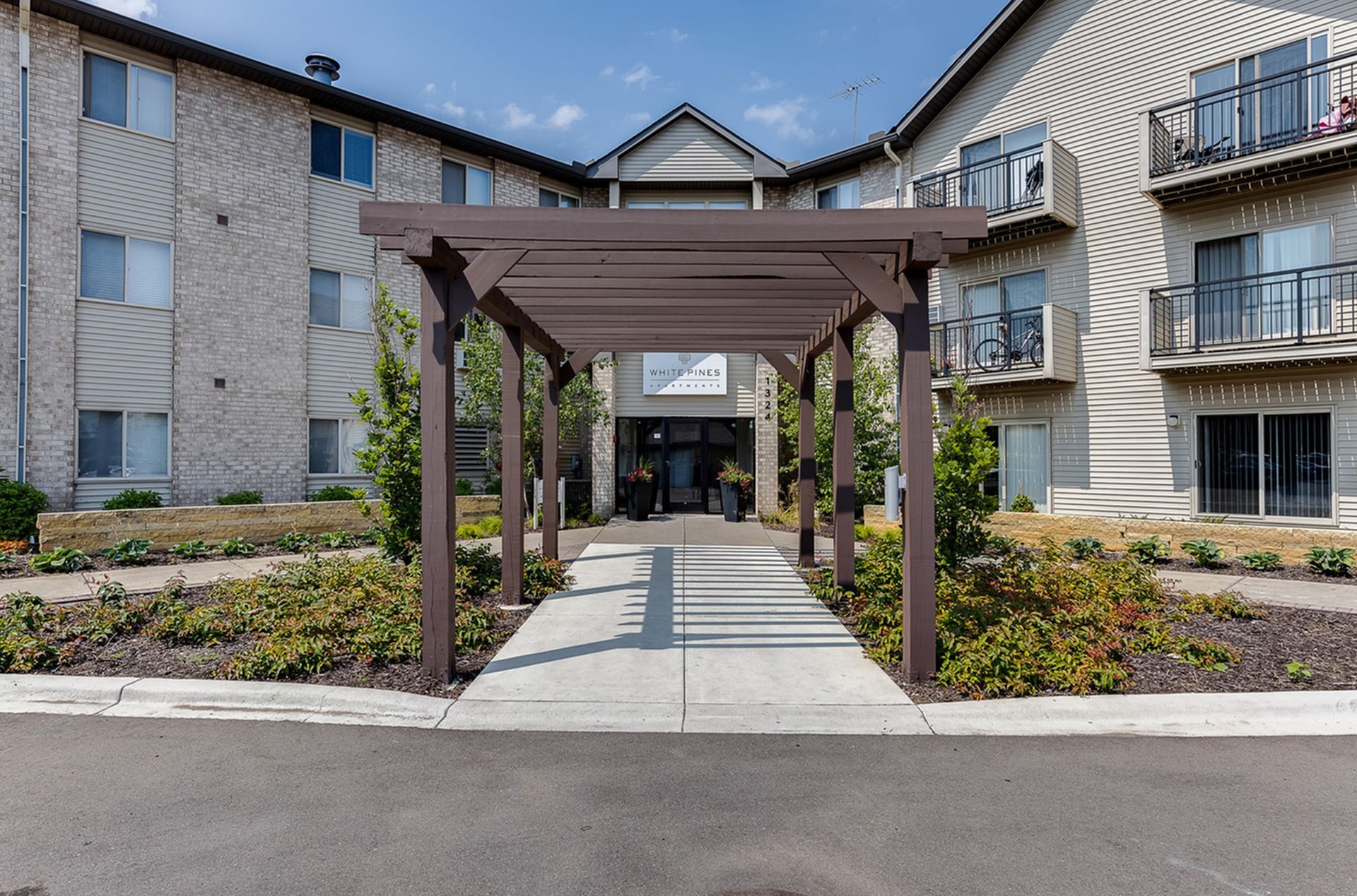 Apartment building entrance with brown pergola walkway, gray sidewalk, and surrounding landscaping.