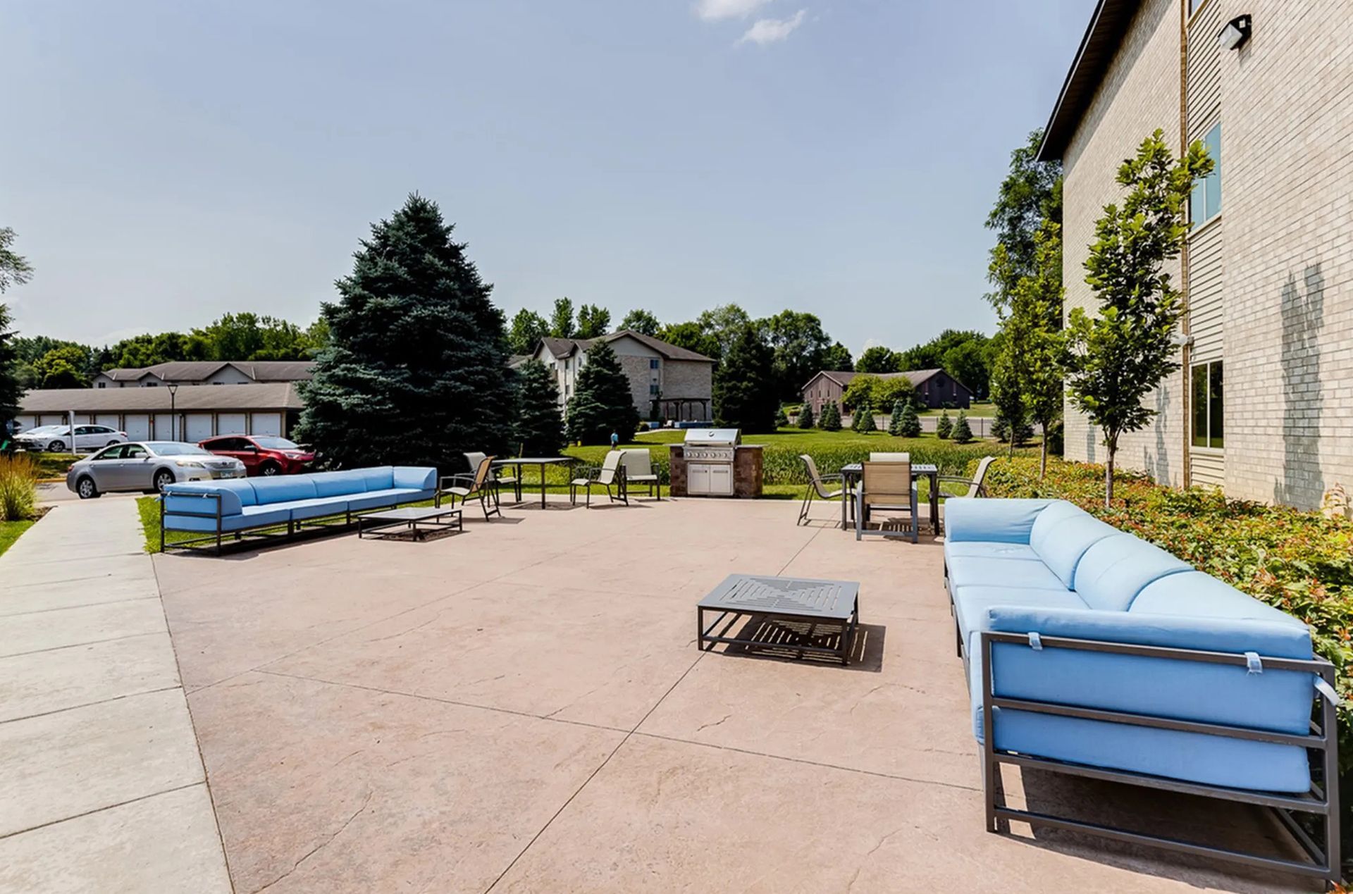 Outdoor patio with blue sofas, tables, chairs, grill, and trees. Cars and a building in the background.