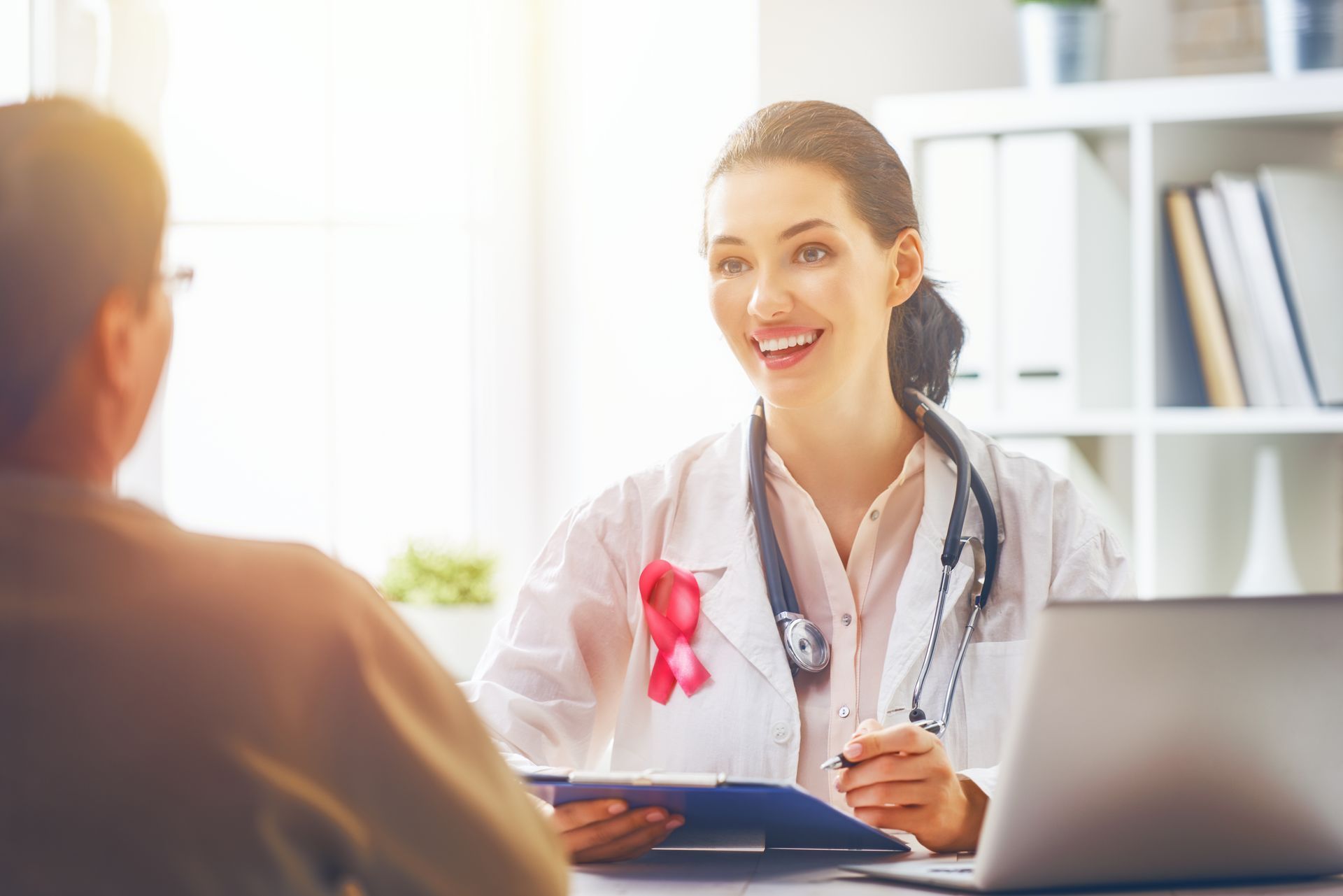 Nurse with stethoscope smiles while talking to a patient in an office; pink ribbon on coat.