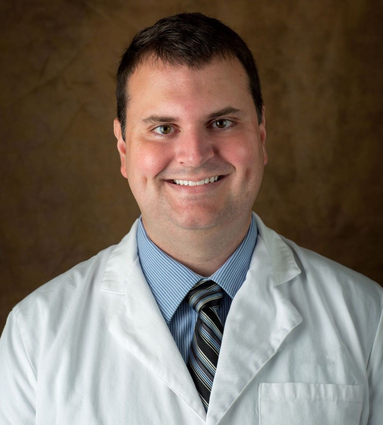 A man in a lab coat and tie smiles for the camera