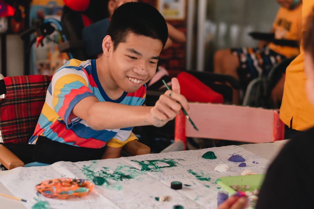 A Young Man is Sitting at a Table Painting With a Brush — Adaptive Care Pty Ltd In Tarneit, VIC