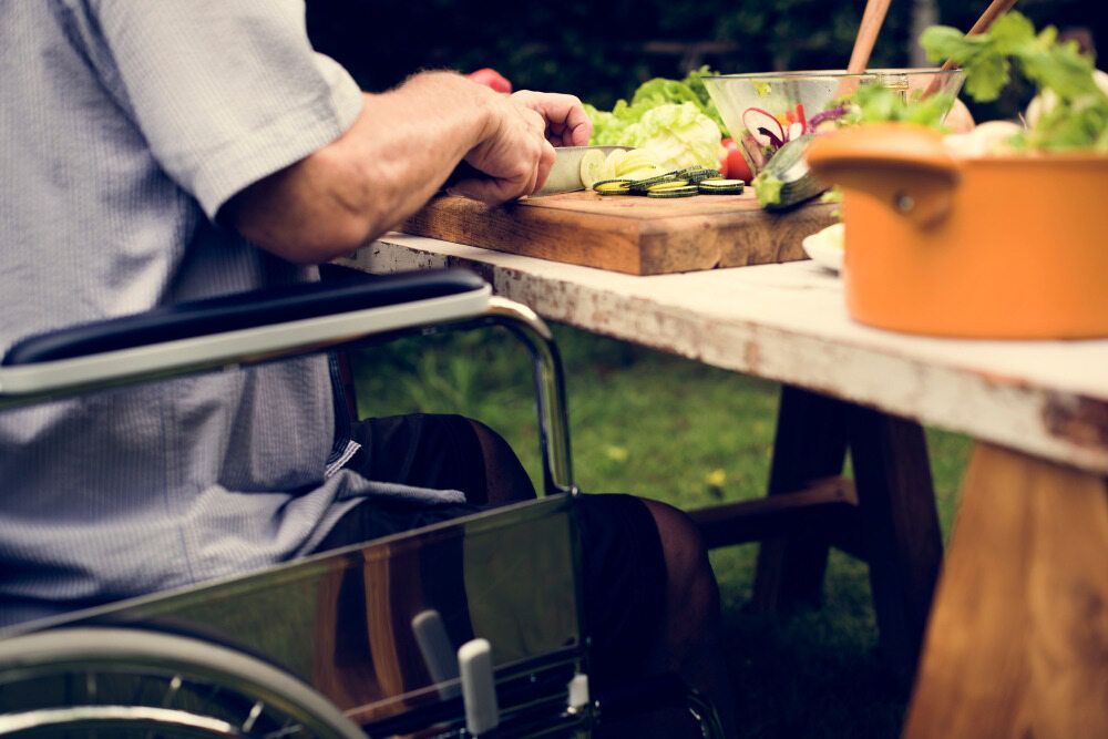 A Person in a Wheelchair Slices Cucumbers on a Wooden Board — Adaptive Care Pty Ltd In Cranbourne, VIC