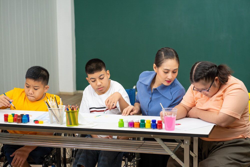 A Teacher Assists Three Children Painting — Adaptive Care Pty Ltd In Preston, VIC