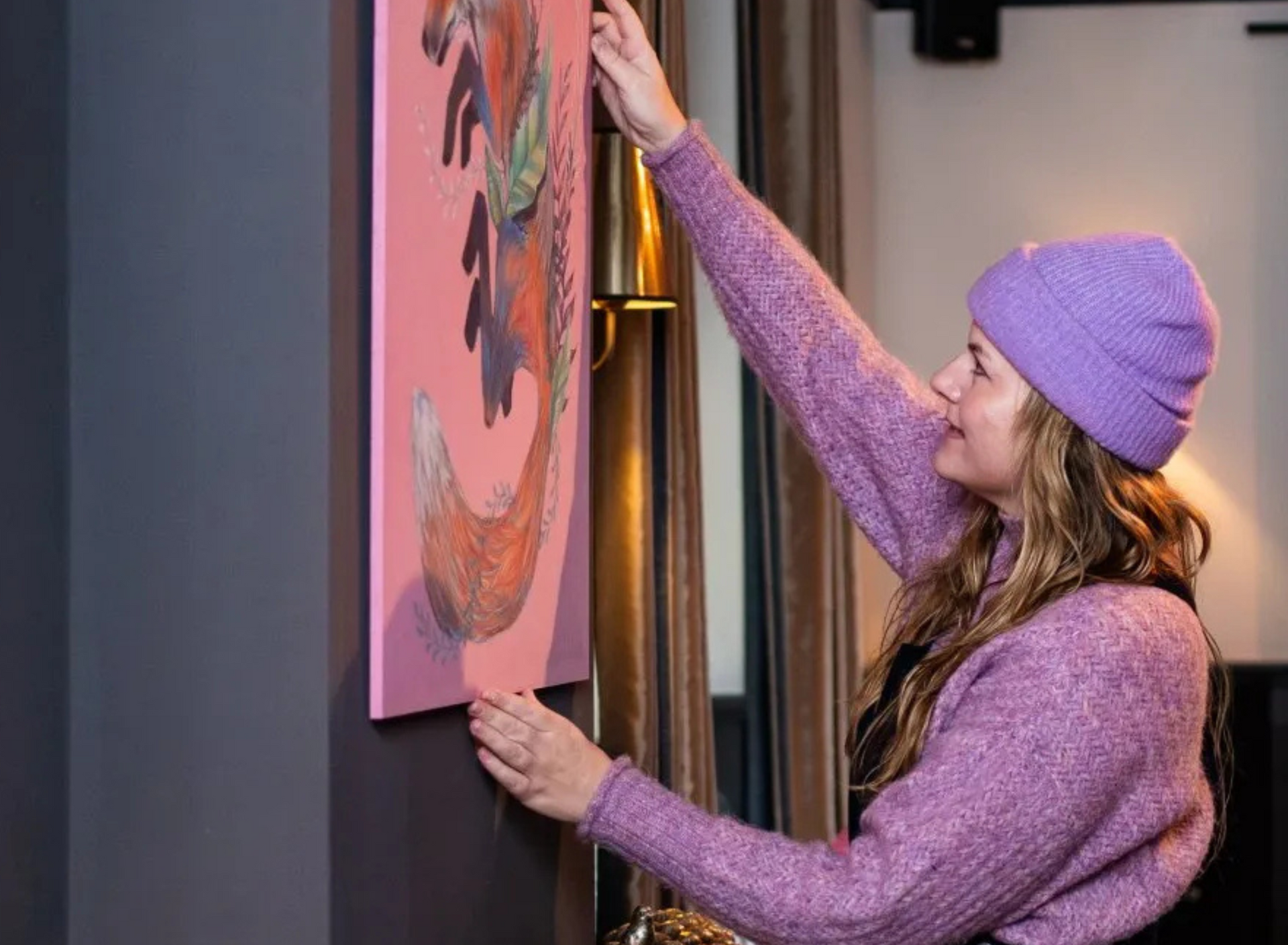 Vrouw in paarse trui en hoed hangt kunstwerk van een vos op tegen een roze achtergrond.