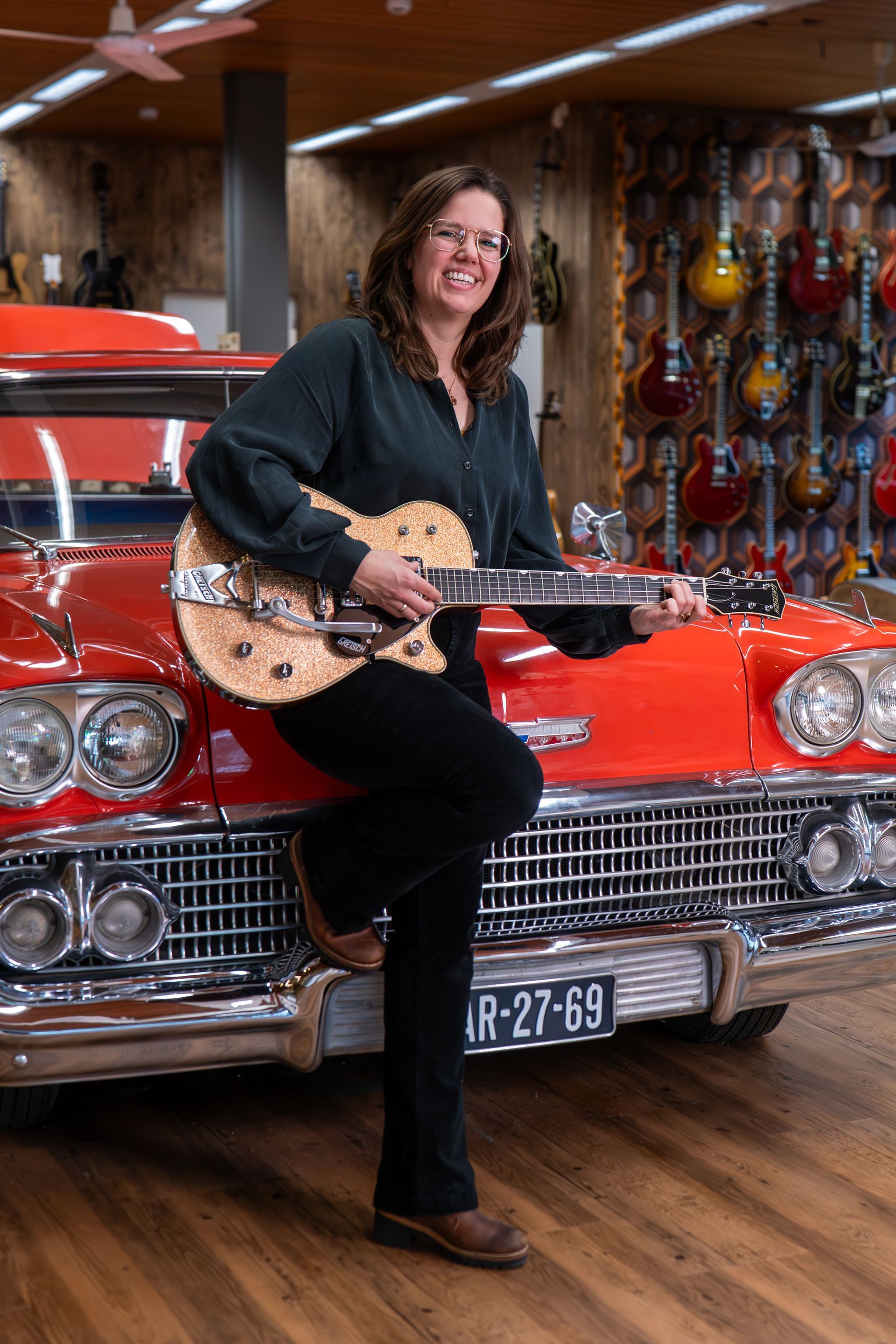 Vrouw met gitaar in de hand, leunend tegen een rode klassieke auto, in een gitaarwinkel.