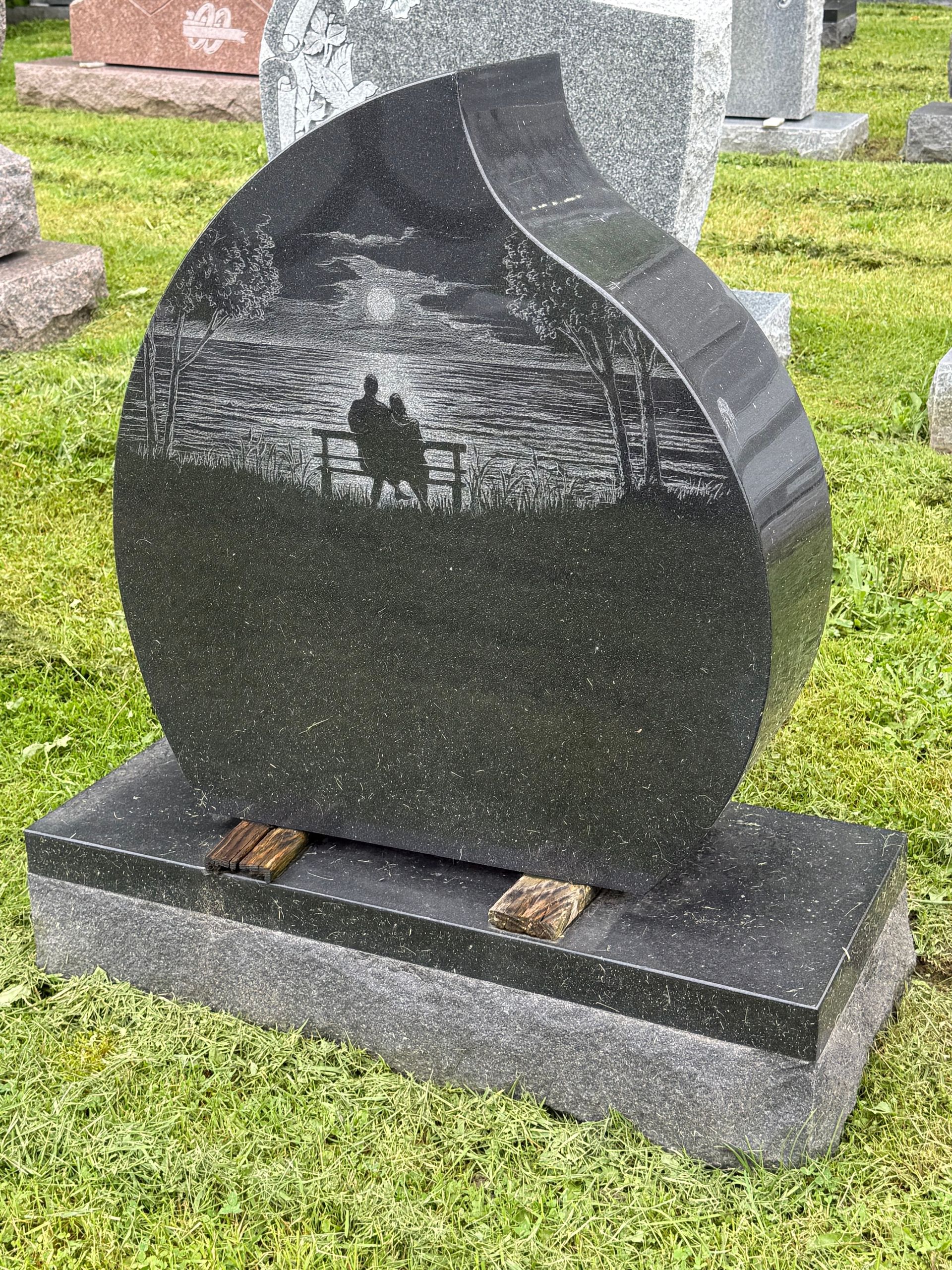 A black gravestone with a silhouette of two people sitting on a bench in a cemetery.