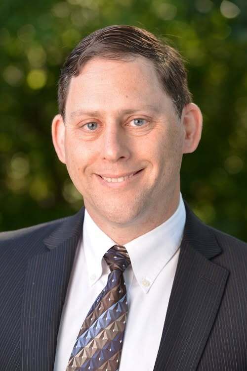 Professional headshot of a man wearing a dark suit, white shirt, and patterned tie against a soft green foliage background.