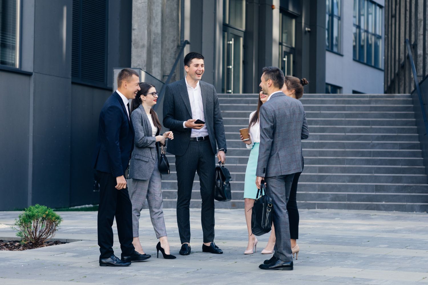 A group of colleagues in business attire stand on a plaza before stairs, laughing and chatting together.