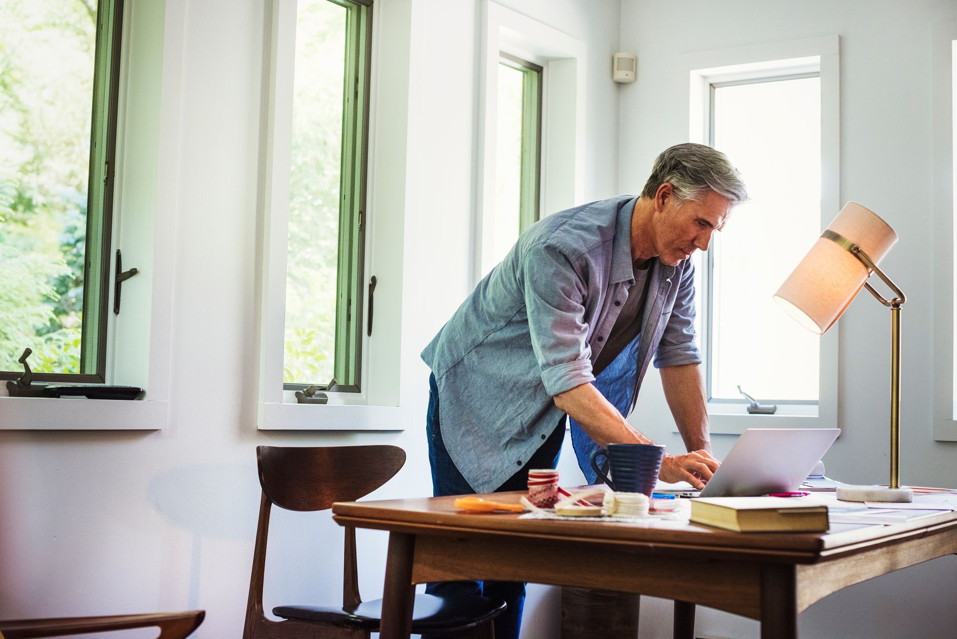 A person in a blue button-down shirt works at a laptop on a wooden desk in a bright, sunlit room.