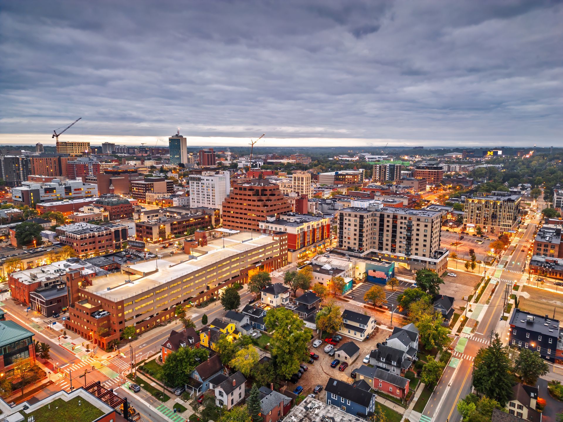 Aerial view of a city at dusk, showing a mix of residential homes, large buildings, and cranes under a cloudy sky.