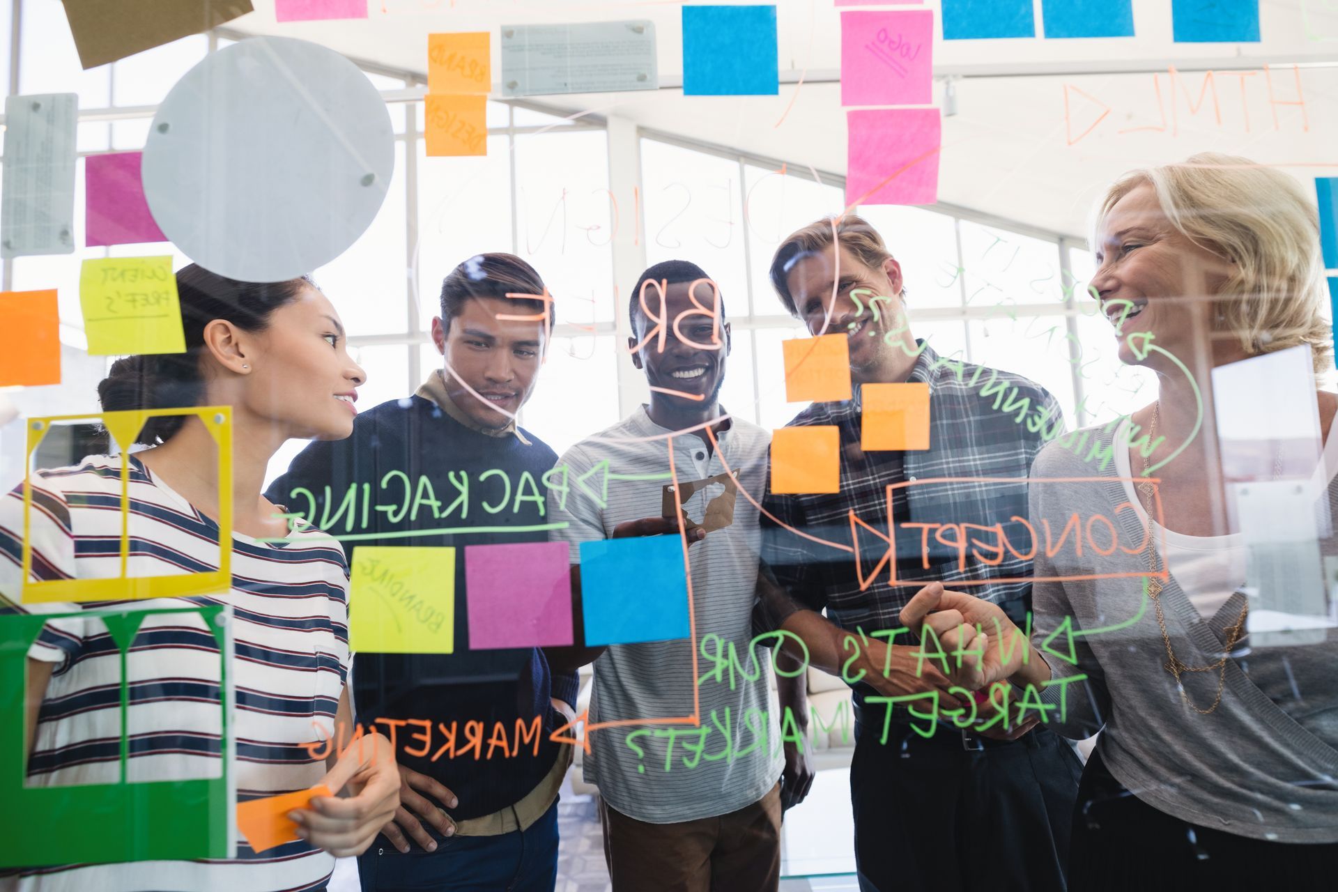 Four diverse colleagues brainstorming while writing notes and drawing on a glass office partition wall.