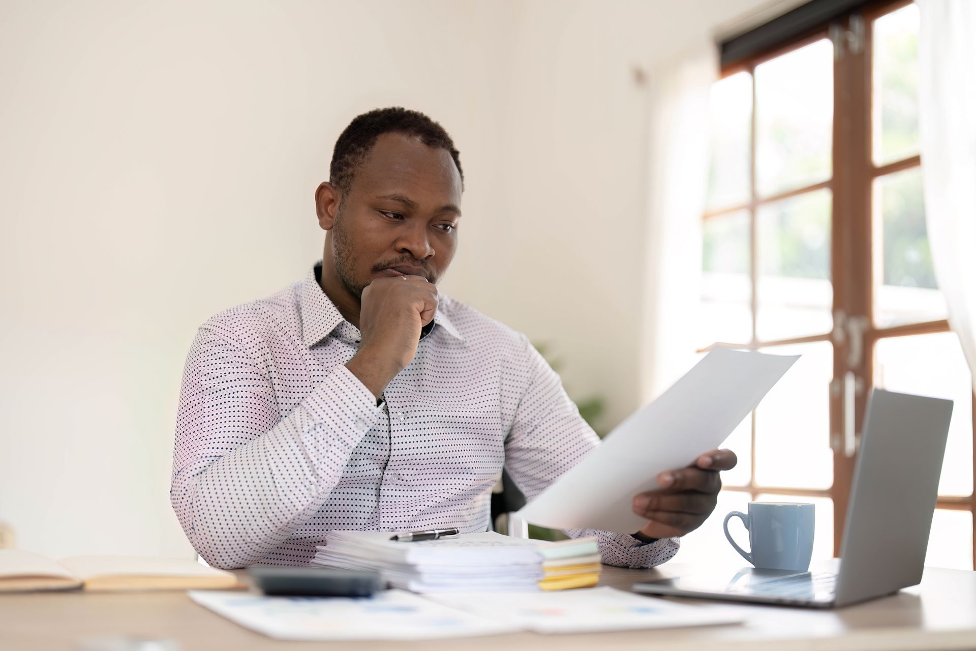A person at a desk looks at documents with a hand to their chin, with a laptop and coffee mug nearby.