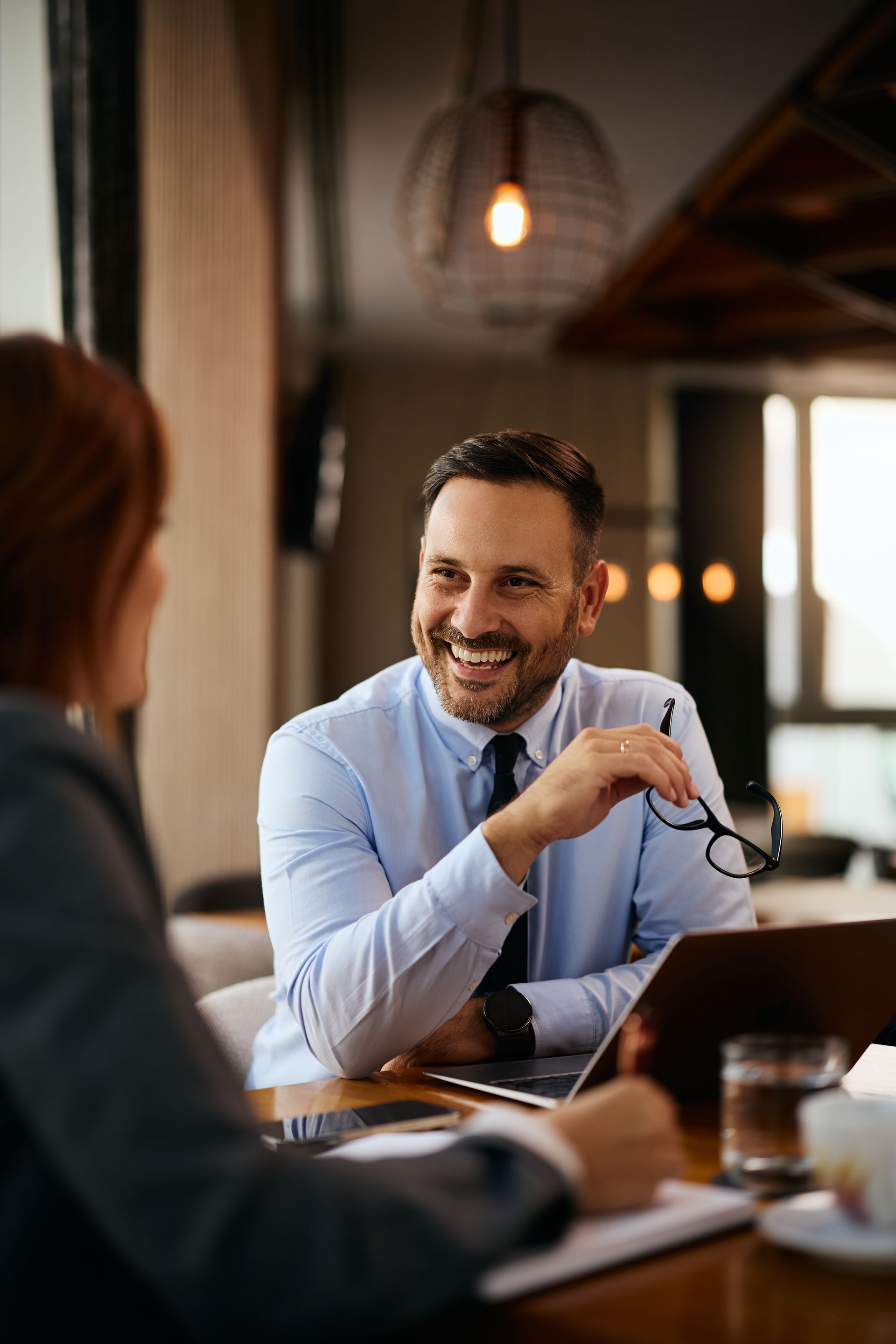 A smiling professional in a blue shirt holds glasses while talking to a colleague across a table in a warm office space.