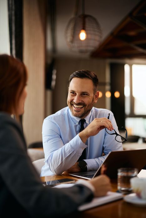 A smiling professional in a blue shirt holds glasses while talking to a colleague across a table in a warm office space.