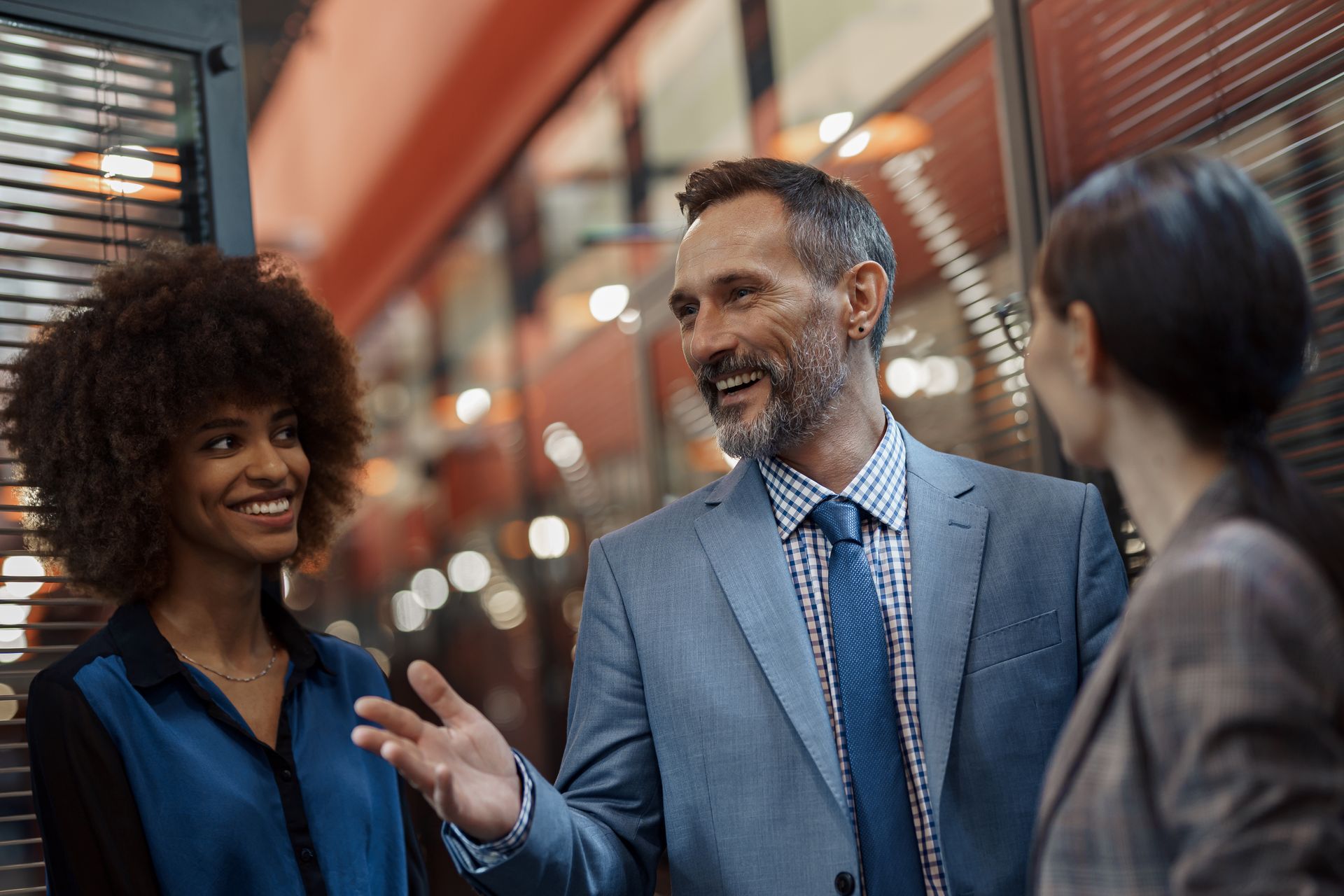 Three professionals in business attire conversing in a bright office hallway, with the center individual gesturing.