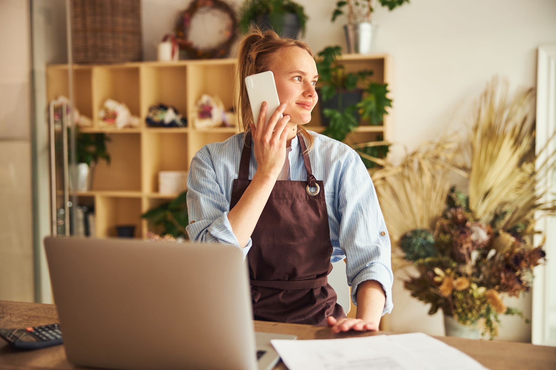 A person in a brown apron talks on a phone while standing at a desk with a laptop, in a shop filled with plants.