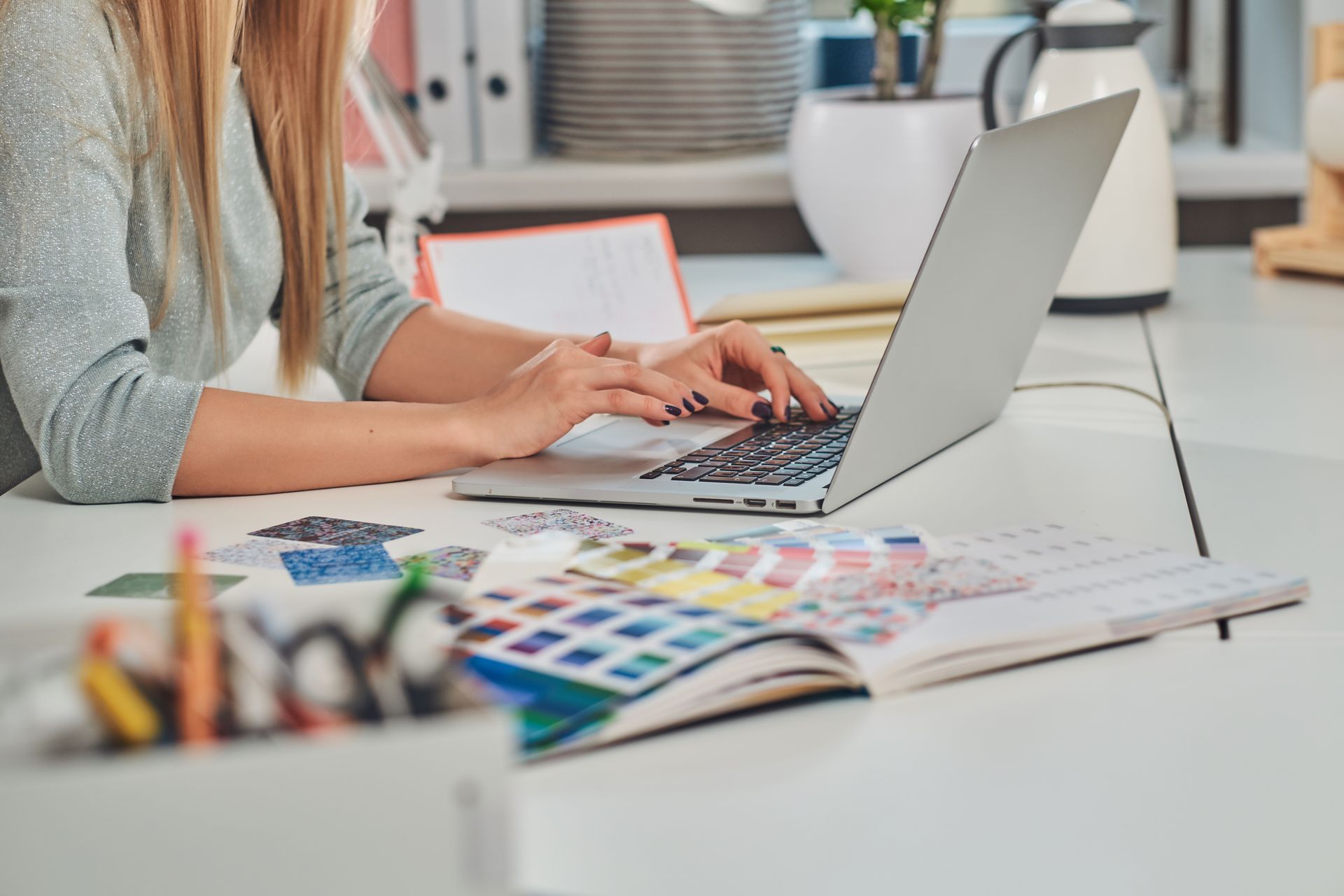 A person typing on a laptop at a desk with color swatches and art supplies in a workspace.