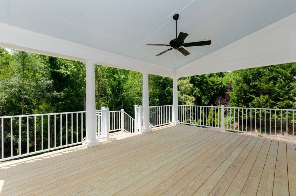 Covered outdoor deck with white railing, ceiling, and columns; black ceiling fan; lush green trees visible.