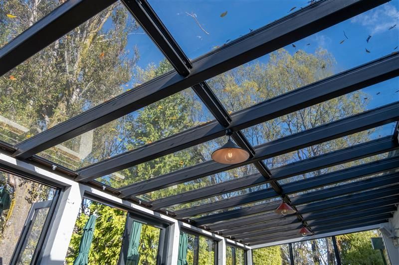 Glass roofed patio with black beams, overlooking trees, and a hanging light.