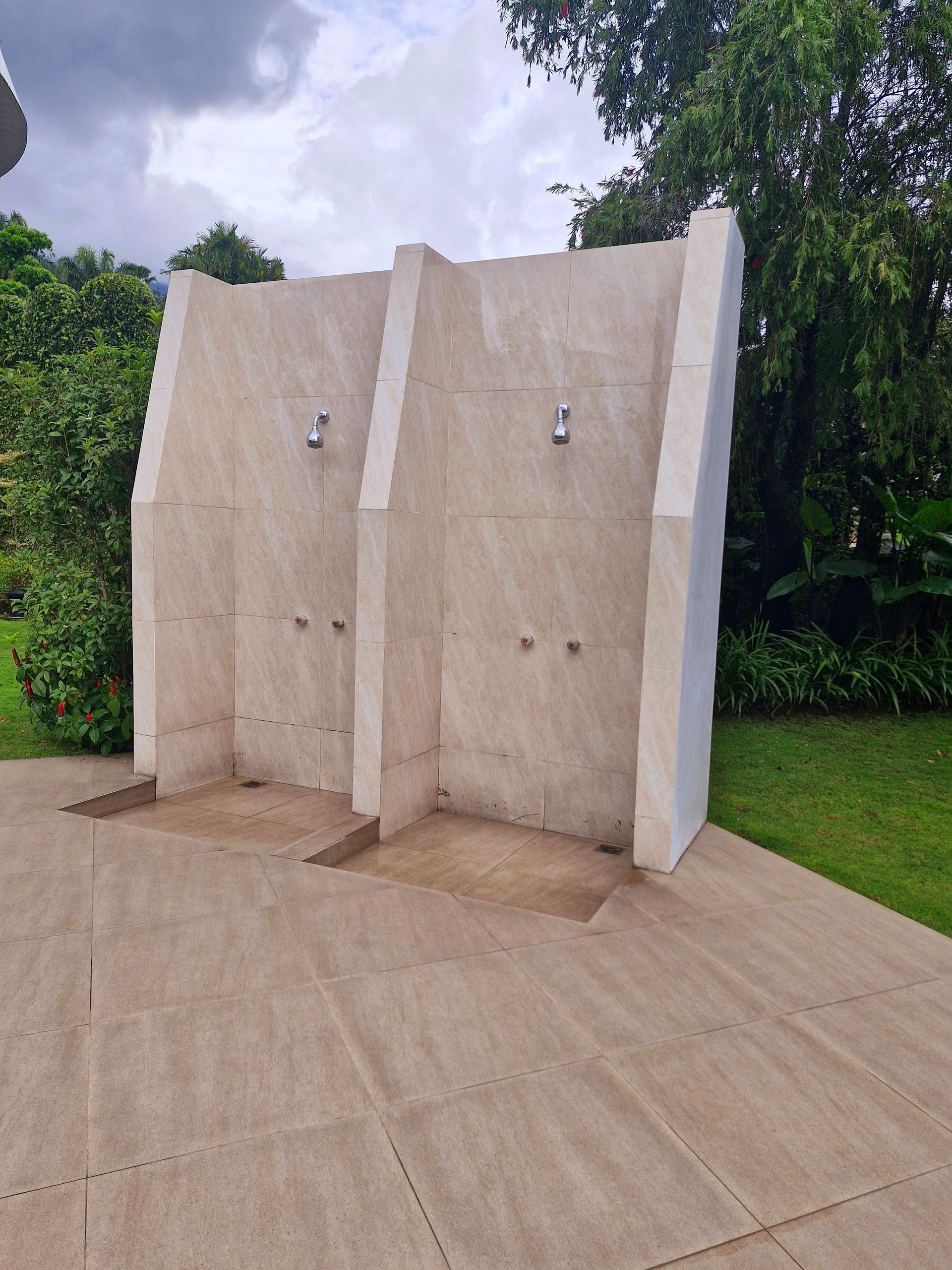 Two outdoor showers with beige tile walls, set on a concrete slab, surrounded by grass and greenery.