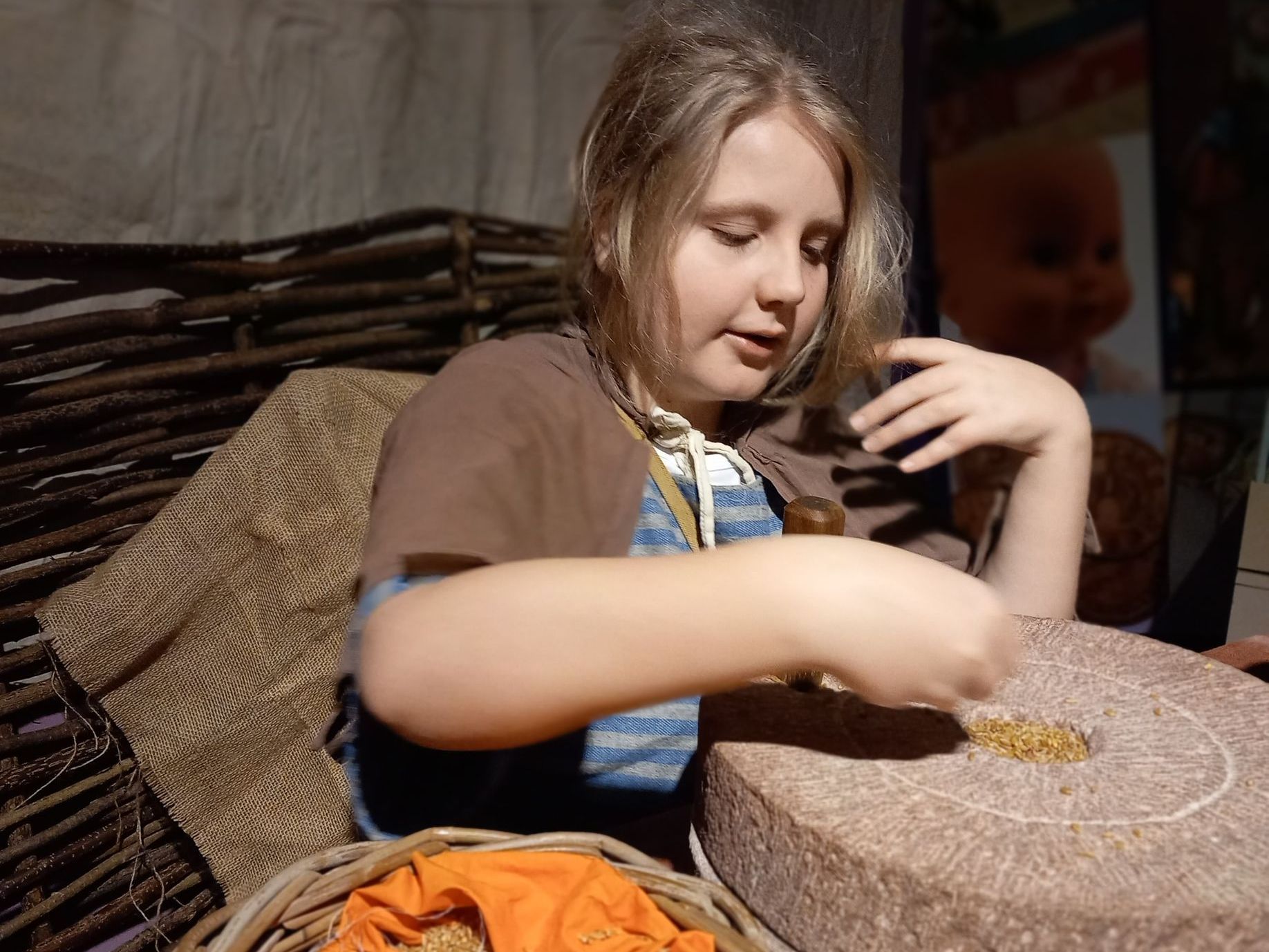 A young boy in a brown cloak sits in front of a woven wall.  He has wheat grain in his hand and is adding it to a circular stone.