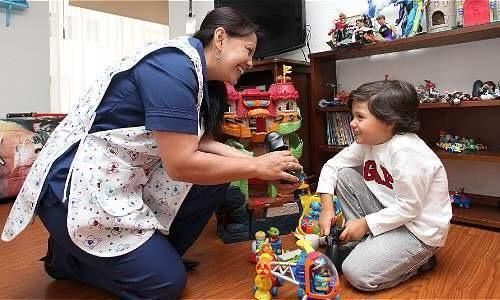 Una cuidadora, arrodillada junto a un niño sentado, le entrega un juguete en una sala de juegos llena de juguetes coloridos.