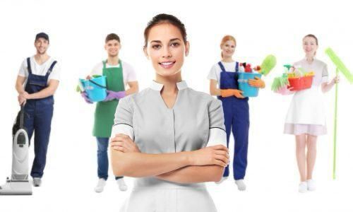 Equipo de limpieza con suministros; mujer sonriente en primer plano, trabajadores detrás sobre fondo blanco.