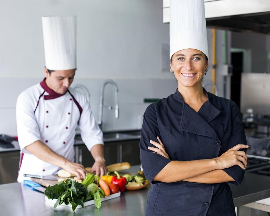 Cocineros con uniformes blancos preparando comida en una luminosa cocina industrial; uno de ellos sonríe a la cámara.