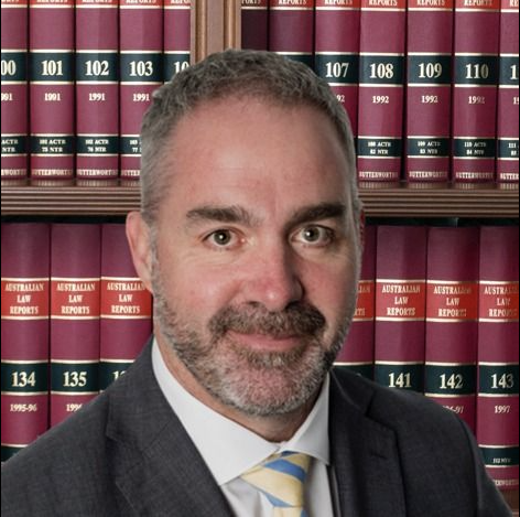 A man in a suit and tie is standing in front of a bookshelf with the number 134 on it