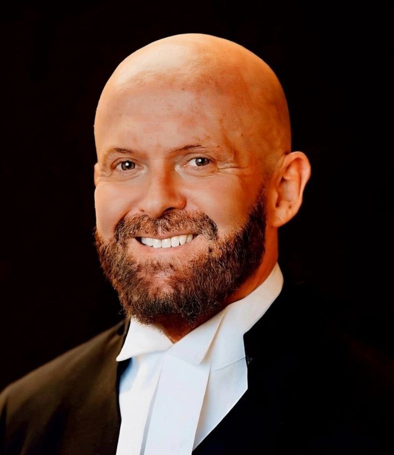 Bald man with beard wearing a black robe and white collar smiling.