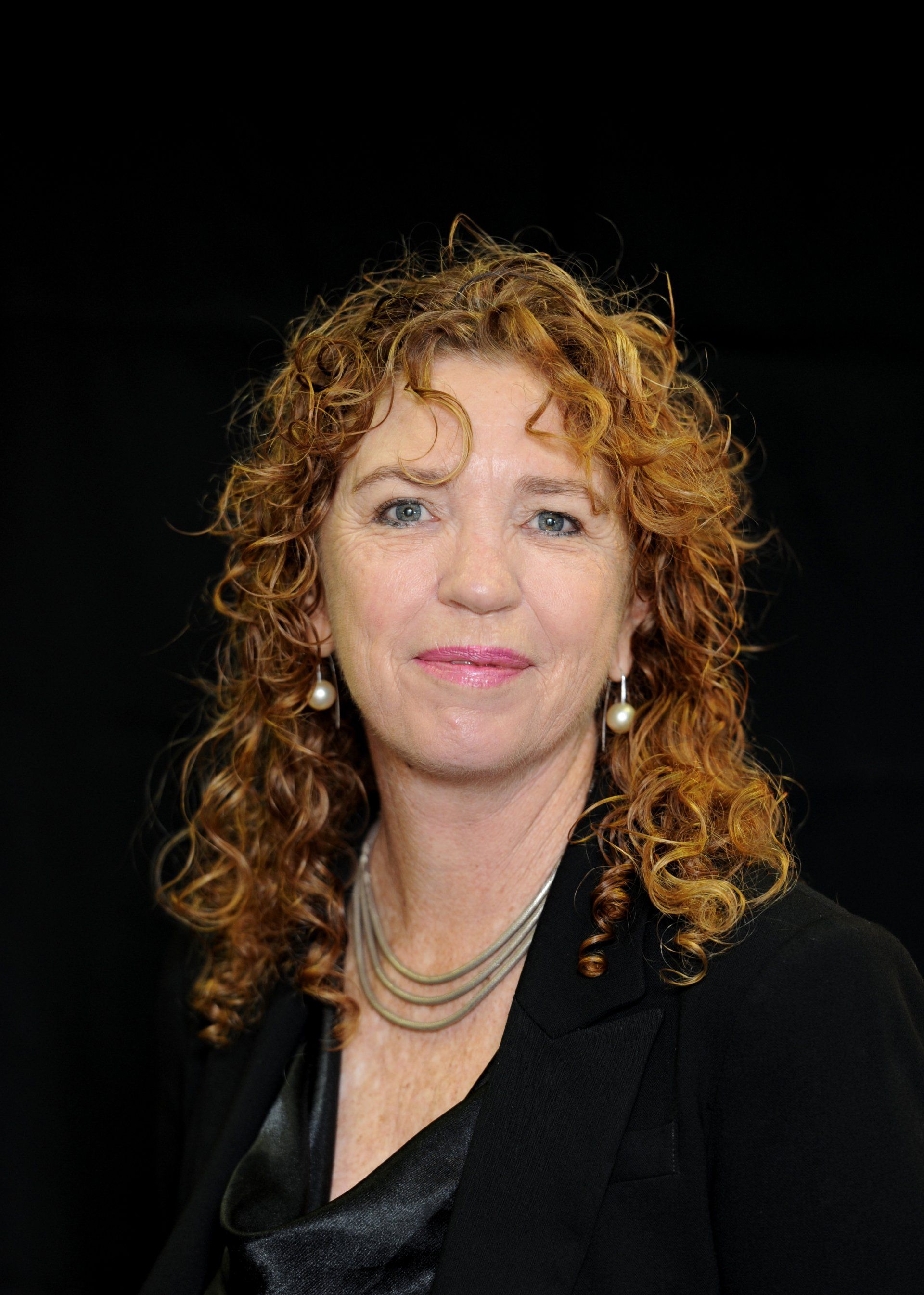 A woman with red curly hair is wearing a black jacket and pearl earrings.
