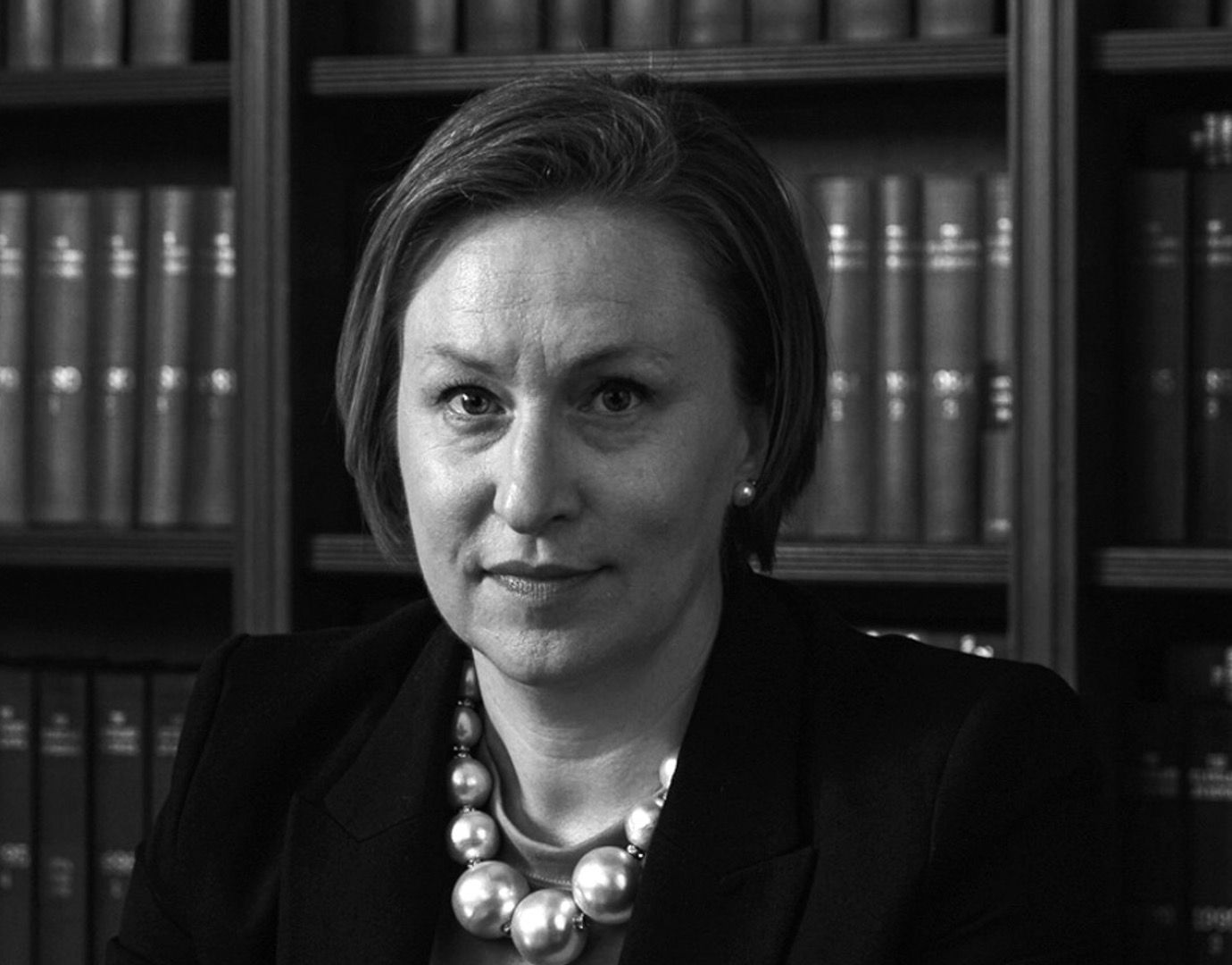 A woman wearing a pearl necklace is sitting in front of a bookshelf.