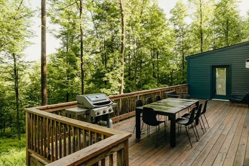 Wooden deck with grill, dining table, and chairs. Green house in background, surrounded by tall trees.