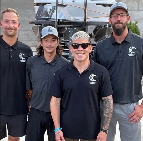 Four people wearing matching black company polo shirts stand in front of a boat on a trailer.