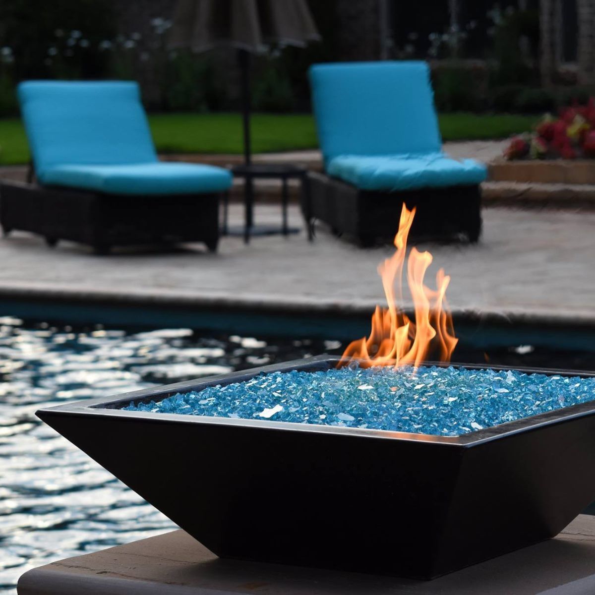 Black fire pit with blue glass and flames, near a pool and lounge chairs.