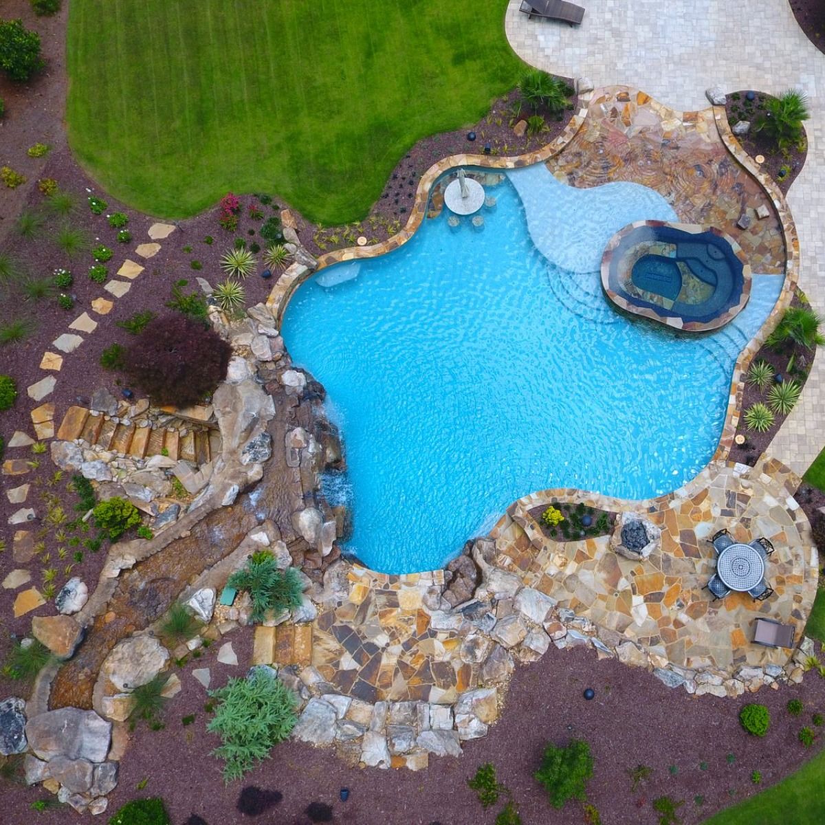 Aerial view of a freeform pool and spa surrounded by stone, landscaping, and green lawn.