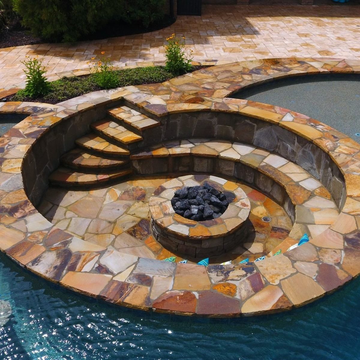 Stone patio with a built-in fire pit, steps, and seats near a pool.