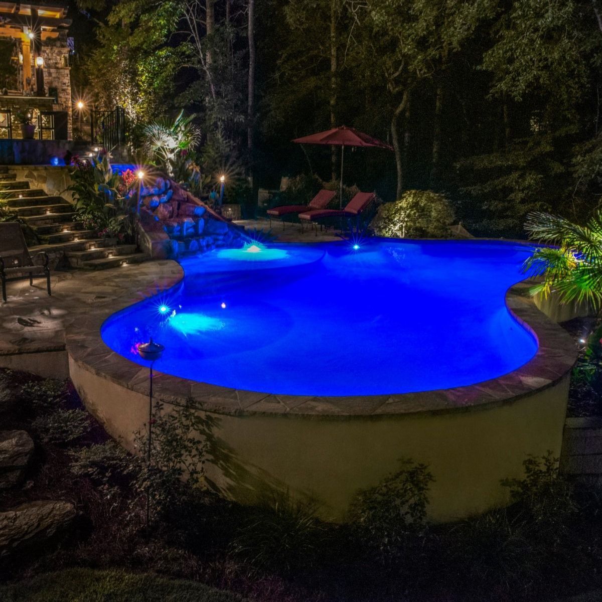Night view of an illuminated blue pool with a stone patio and surrounding trees.