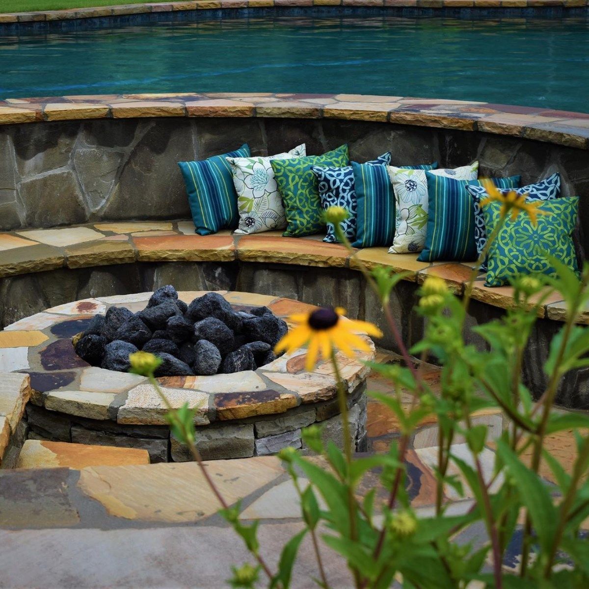 Stone fire pit with built-in seating, pillows, and pool in background.