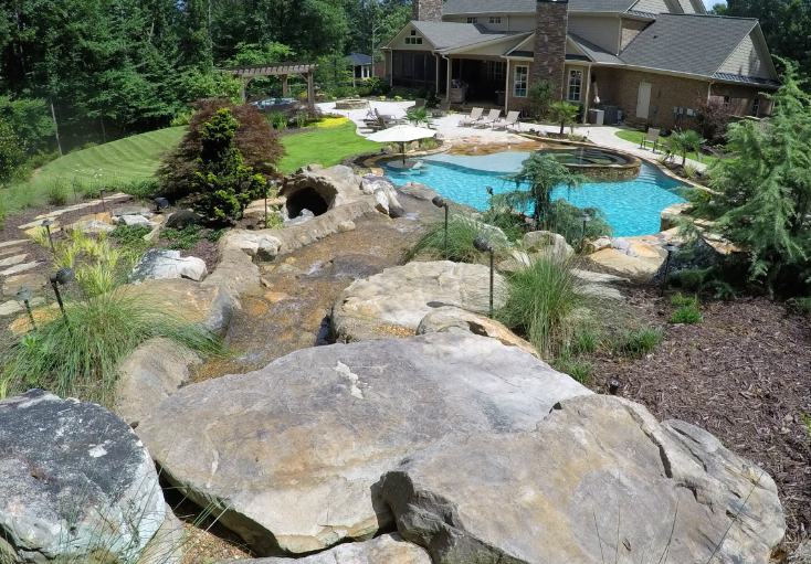 Backyard with pool, waterfall, landscaping, and large house in the background.