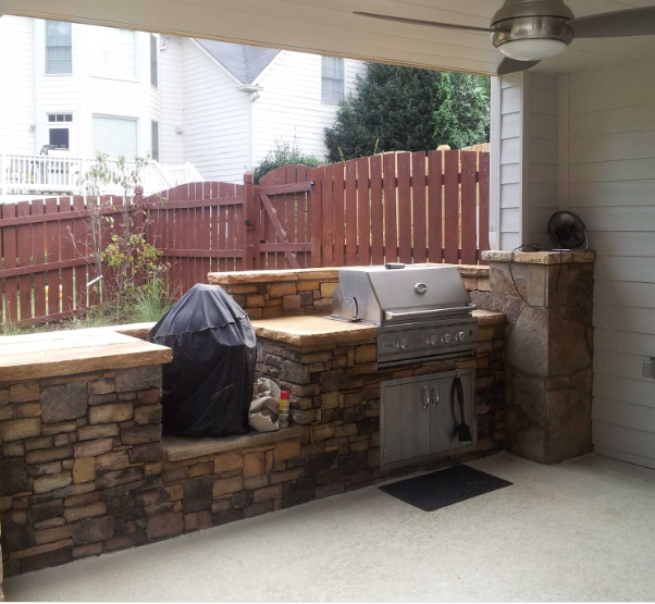 Outdoor kitchen with stone facade, grill, smoker, and wooden fence in the background.