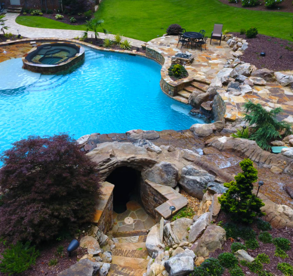 Swimming pool with a cave entrance, waterfall, and hot tub surrounded by rocks and greenery.