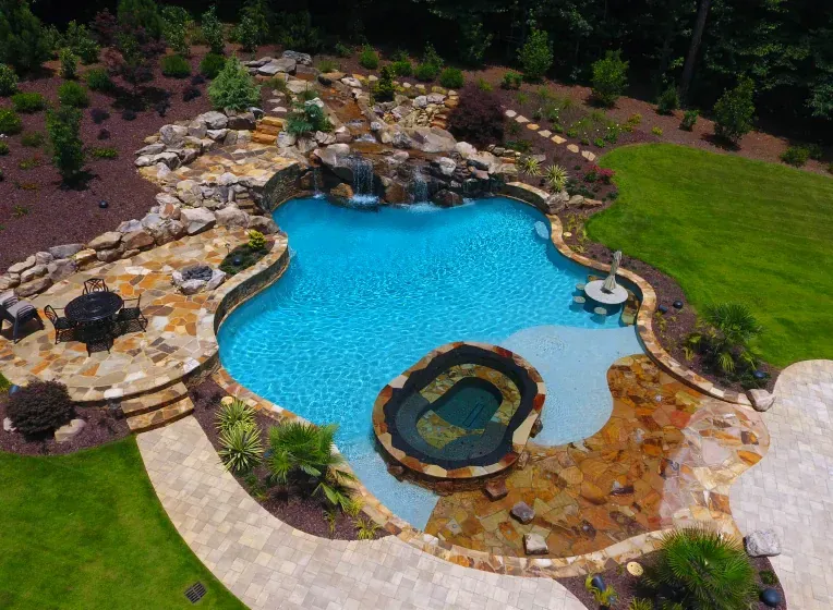 Aerial view of a luxury backyard pool with a waterfall, hot tub, and stone patio. Turquoise water.