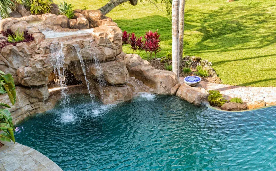 Waterfall cascading into a turquoise pool surrounded by rocks, greenery, and lawn.