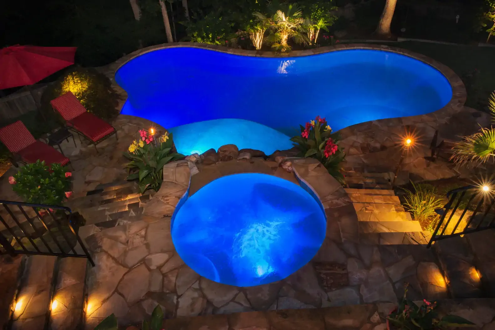 Nighttime view of a blue-lit pool and spa surrounded by stonework and landscaping.