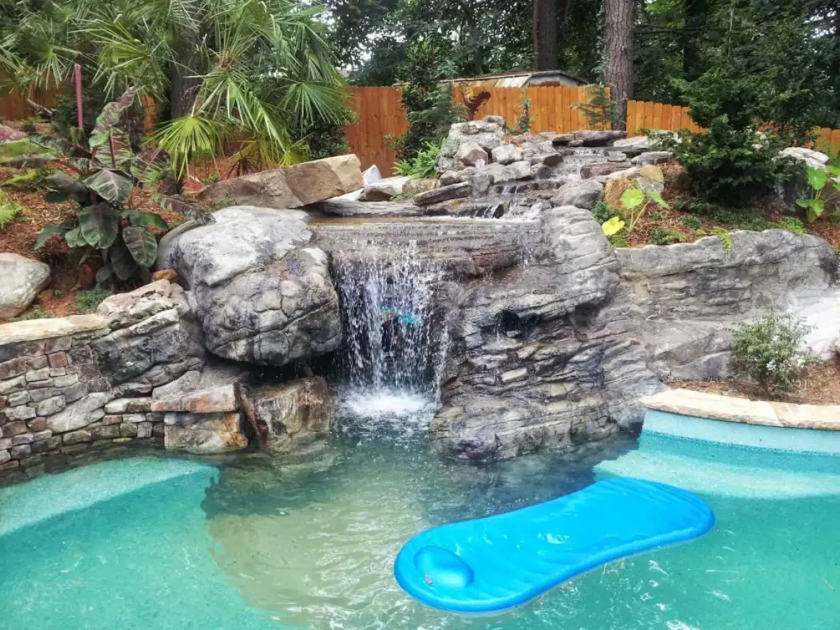 Waterfall cascading into a blue-green swimming pool; a blue floating pad sits in the pool.