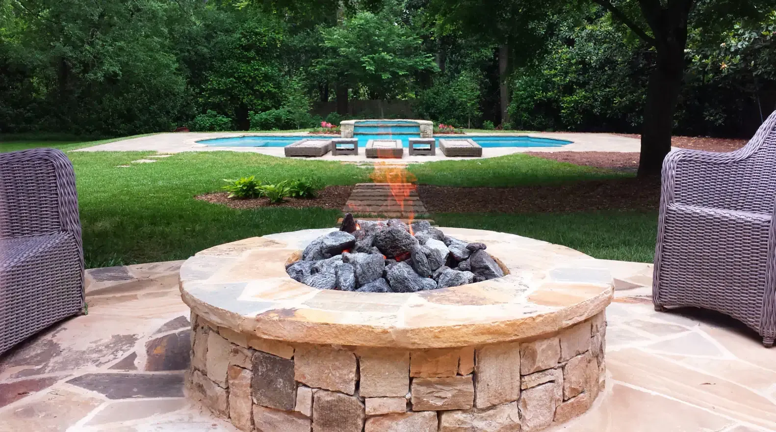 Fire pit on a stone patio, with a pool and chairs in a backyard.