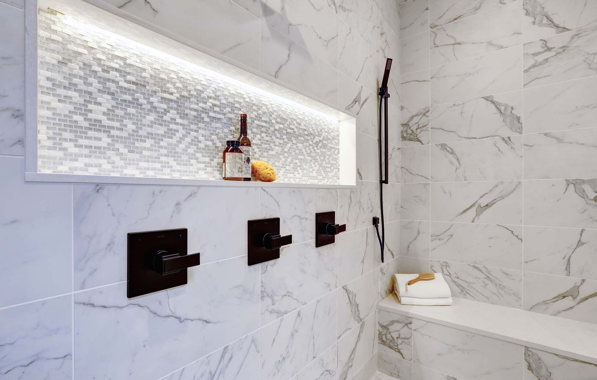 A bathroom with marble tiles , a shower , a bench and a shelf.